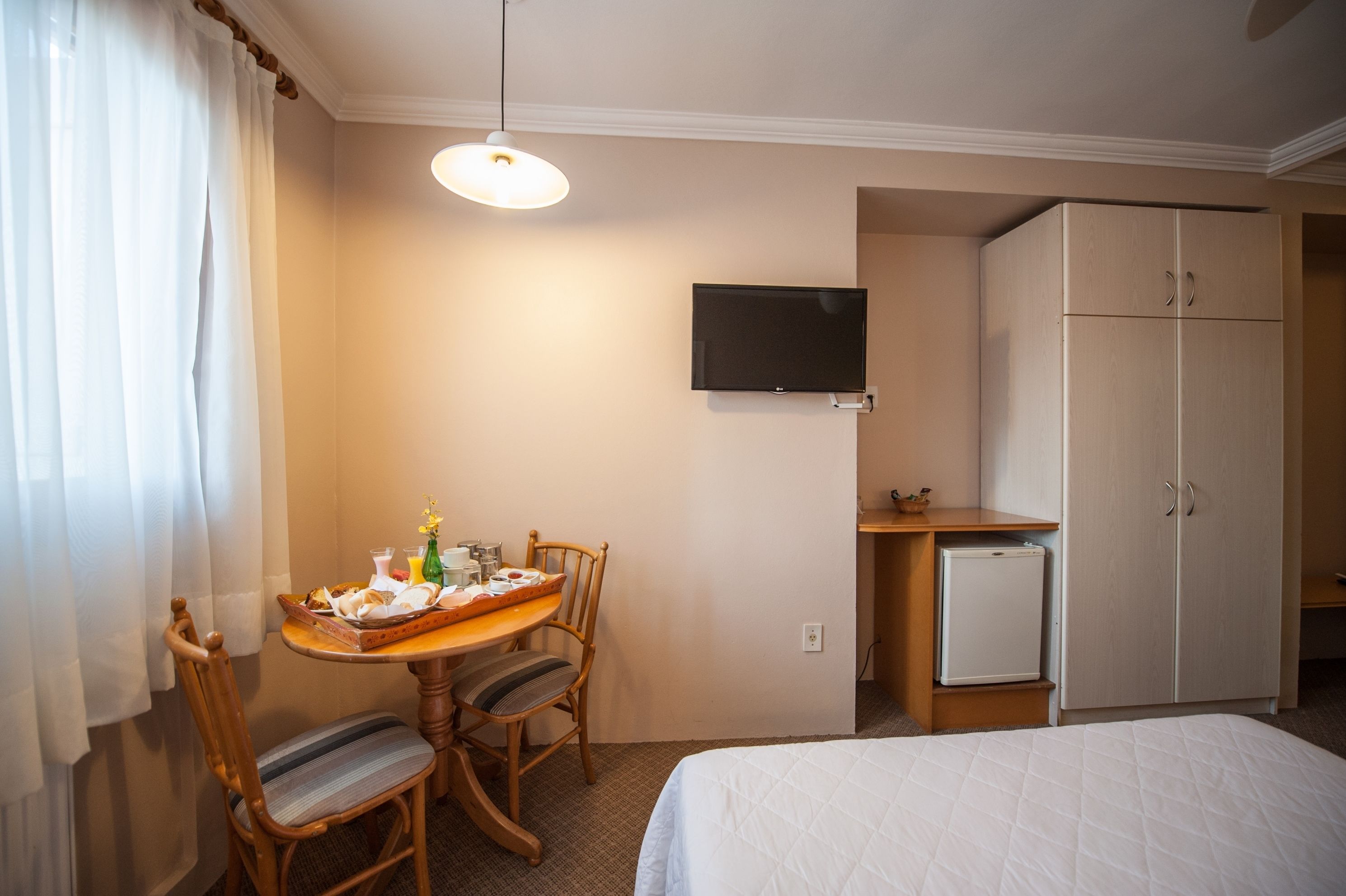 Habitación de hotel con cama, escritorio, silla, televisión, armario y refrigerador pequeño.