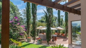 Un espacio al aire libre cubierto con un banco curvo y sombrillas, rodeado de árboles altos y plantas en macetas.