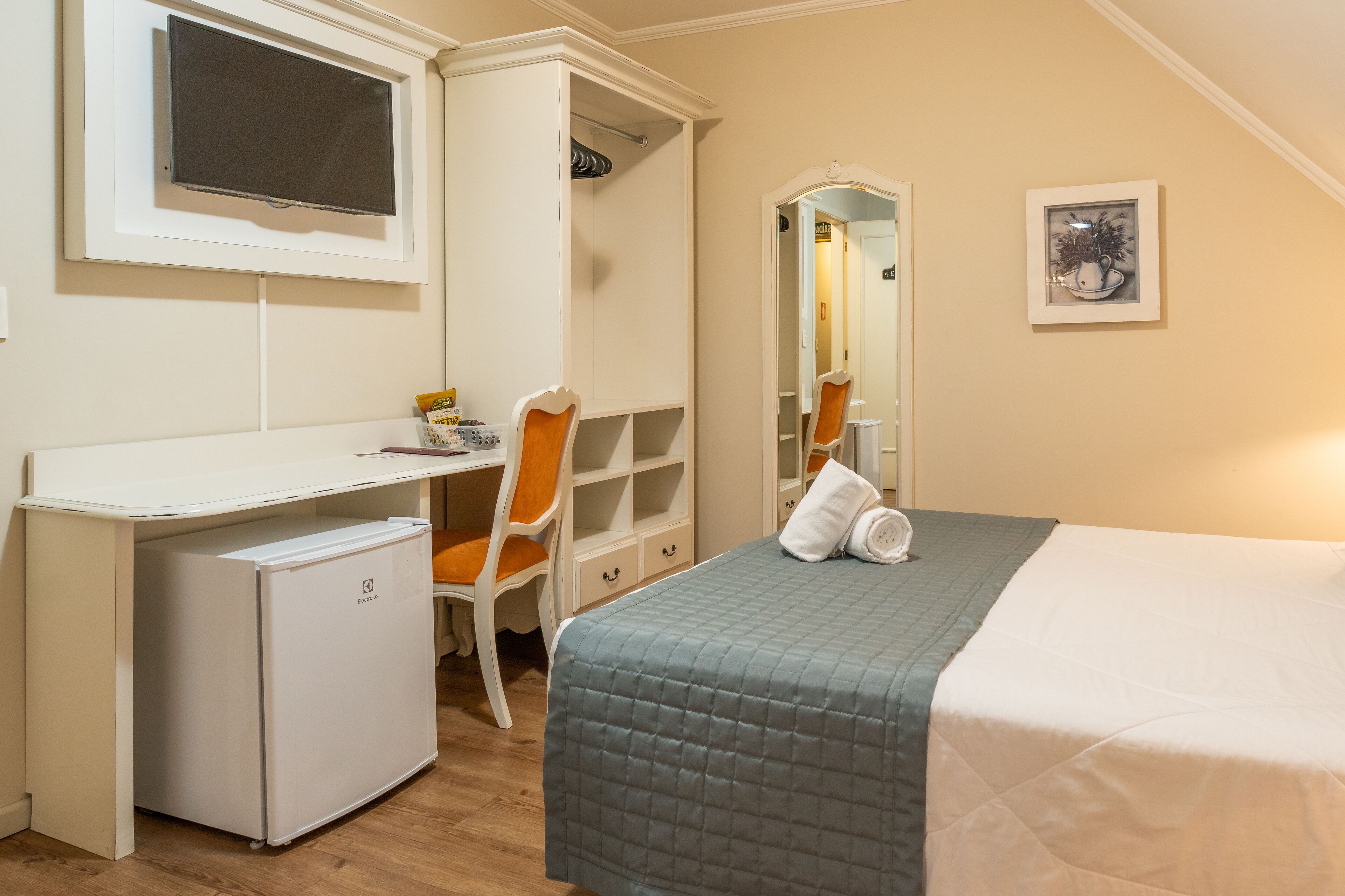 A bedroom with a bed, a desk, a chair, a small refrigerator, a mirror, and a framed picture on the wall.