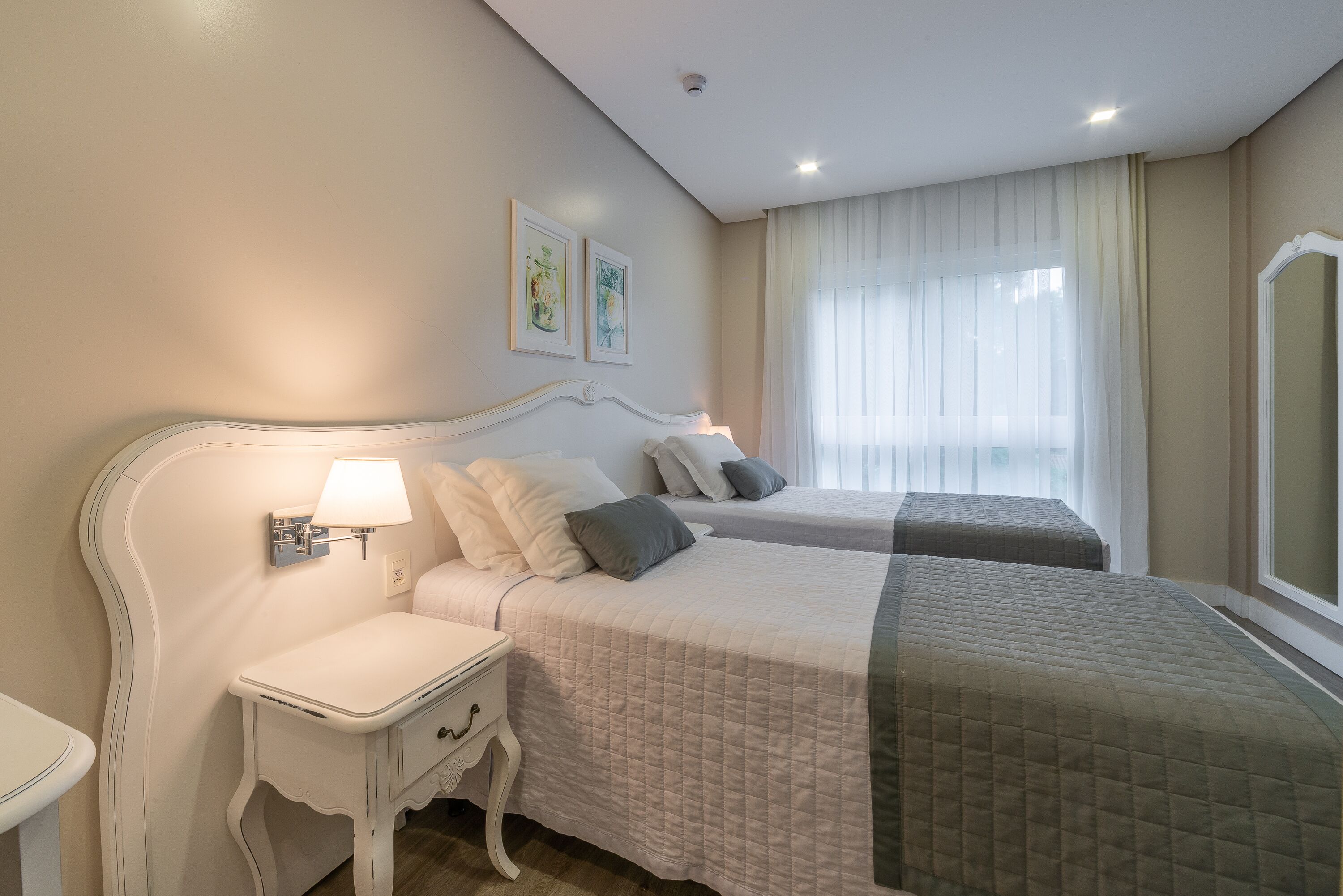 Habitación de hotel con dos camas, una mesita de noche, una lámpara, una ventana con cortinas y dos cuadros en la pared.