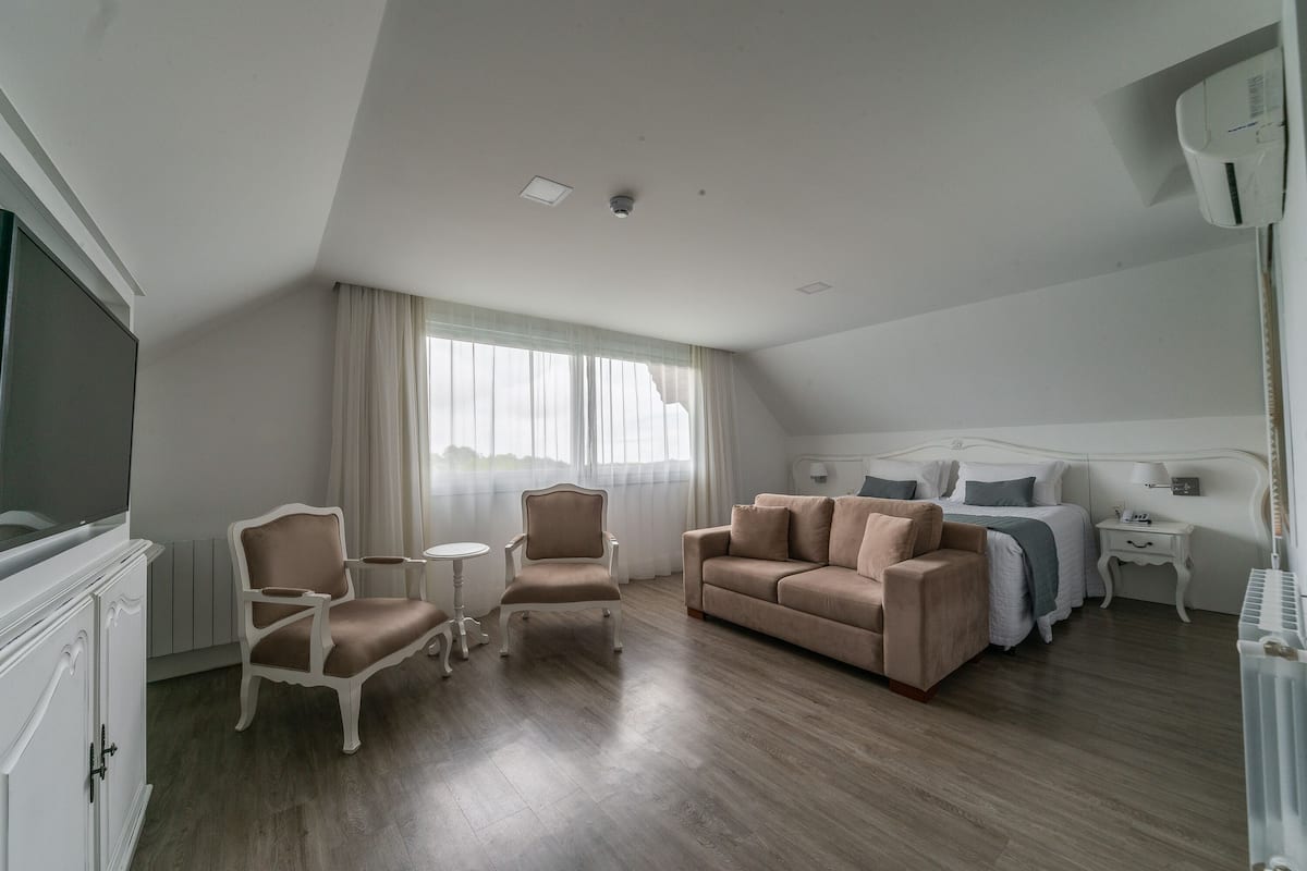 A hotel room with a beige sofa, two armchairs, a small round table, a bed with a white headboard, a nightstand, and a radiator.