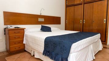 Un dormitorio con cabecera de madera, cama con ropa blanca y una manta azul, mesita de noche con teléfono y un armario de madera.