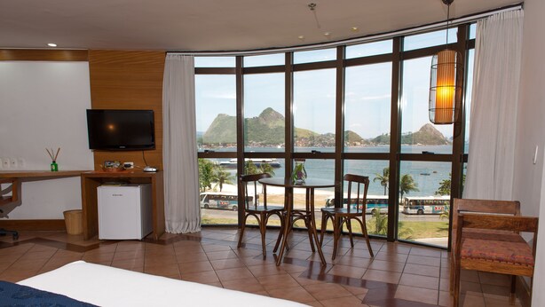 A hotel room with a bed, a desk with a TV, a small fridge, a table with chairs, and a large window with a view of the sea and mountains. - Solar do Amanhecer (Niteroi)