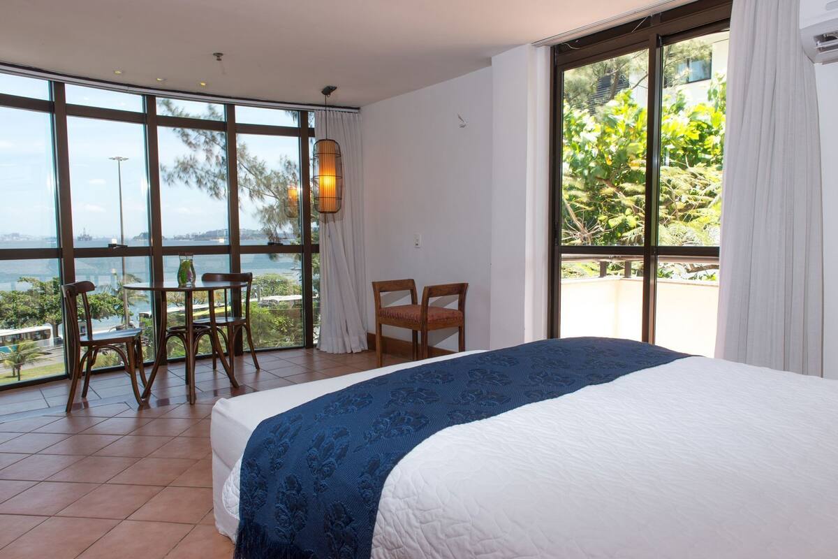 A hotel room with a large bed, a dining table, two chairs, and a view of the sea.
