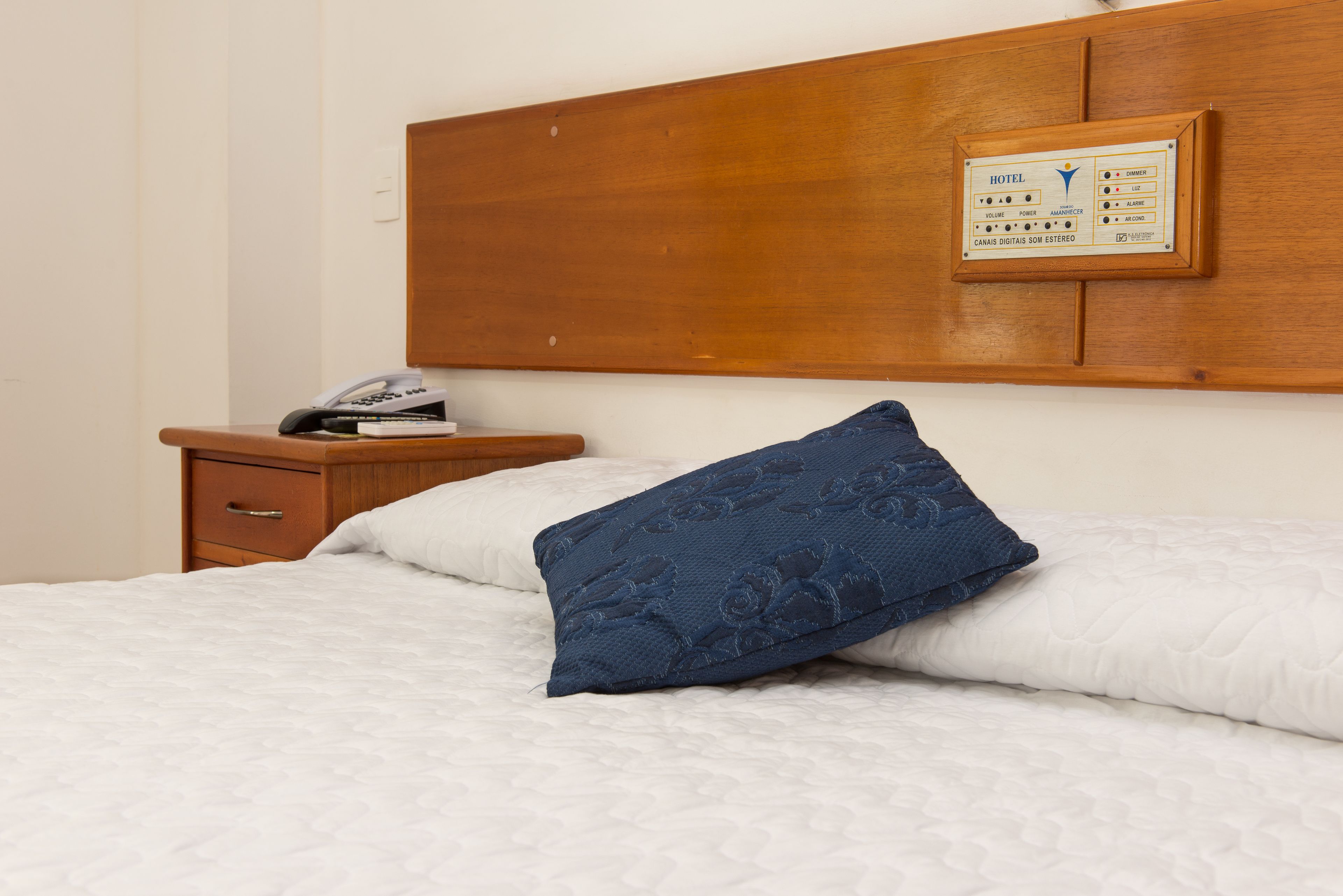 Ein Hotelzimmer mit einem hölzernen Kopfteil, einem Nachttisch mit Telefon und Büchern sowie einem blau gemusterten Kissen auf dem Bett.
