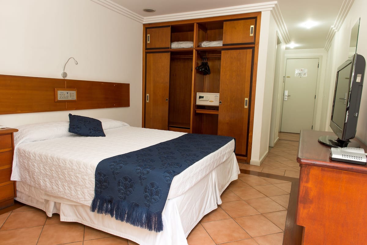 A bedroom with a bed, a wooden wardrobe, a desk with a computer, and a bathroom door.
