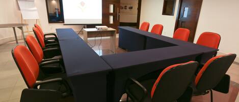 Un salón de conferencias con una larga mesa, varias sillas, una pantalla de proyector y una pizarra.