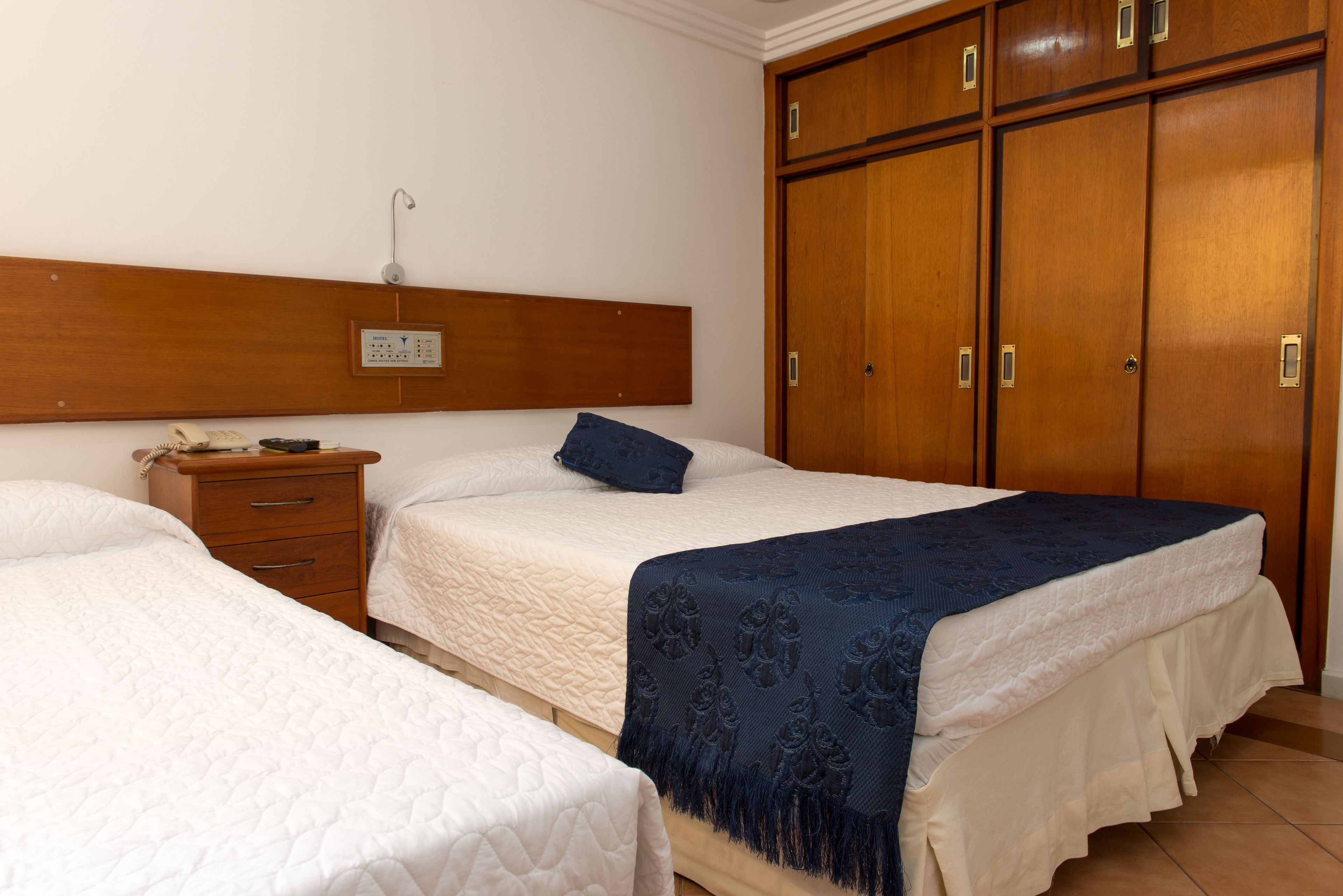Habitación de hotel con dos camas, un armario de madera, una mesita de noche con lámpara y un panel de control en la pared.
