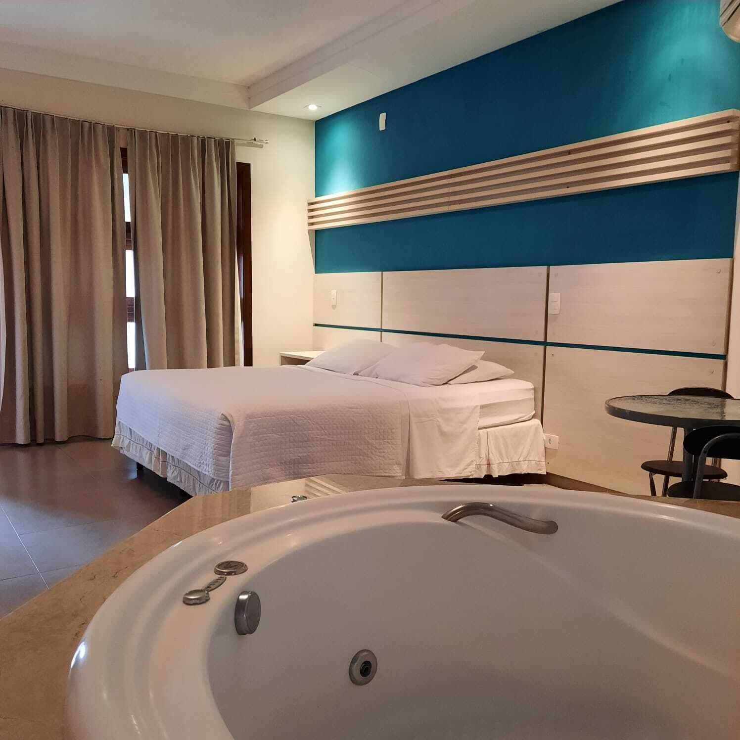Camera d'albergo con una grande vasca, un letto con lenzuola bianche, una parete azzurra e una finestra con tende.