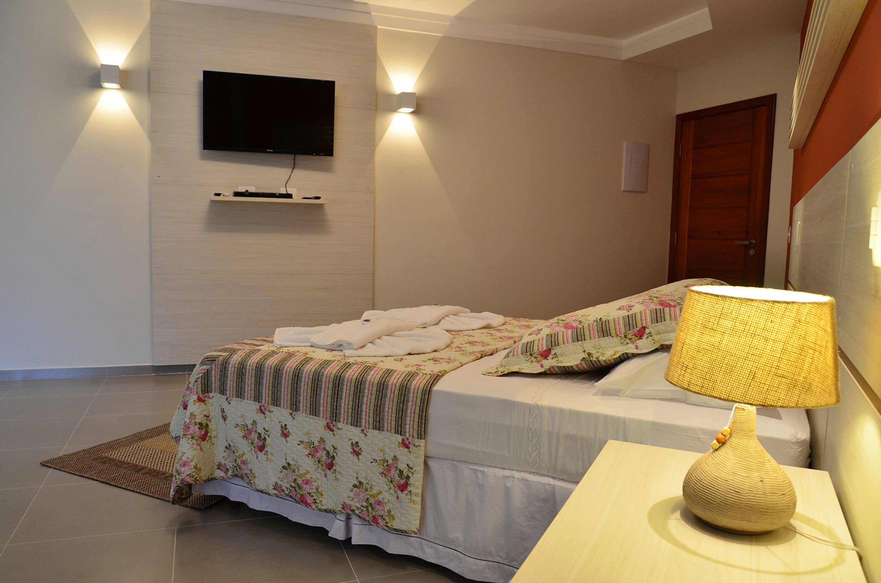A hotel room with a bed, a bedside lamp, a flat-screen TV mounted on the wall, and a wooden door.