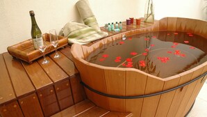 Une baignoire en bois, avec des fleurs rouges flottant sur l’eau, une bouteille de champagne et deux verres posés sur un banc en bois.