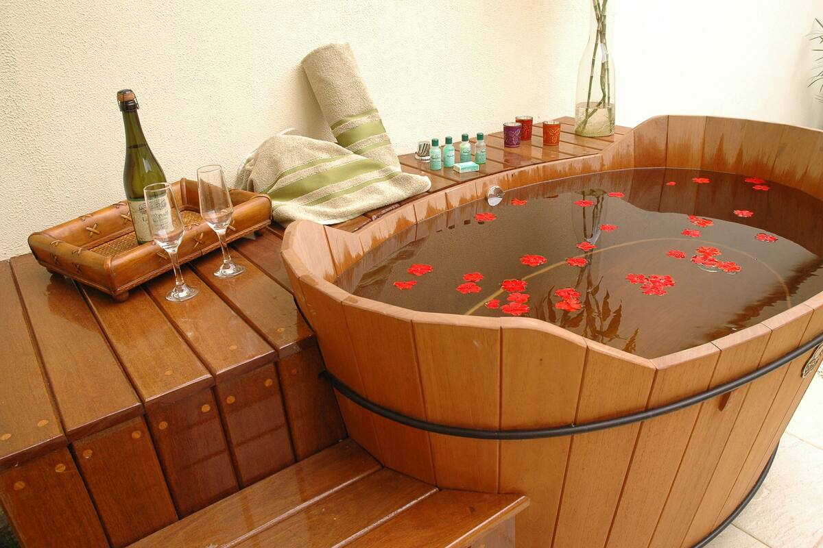 Una bañera de madera con flores rojas flotando en el agua, una botella de champán y dos copas sobre un banco de madera.