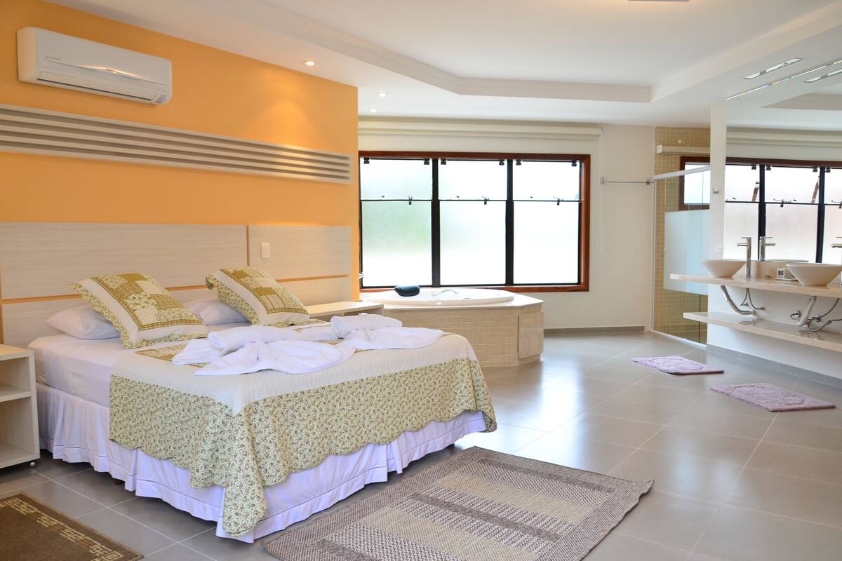 Una habitación de hotel moderna con una cama grande, una mesita de noche redonda, una bañera integrada y un ventanal que proporciona luz natural.