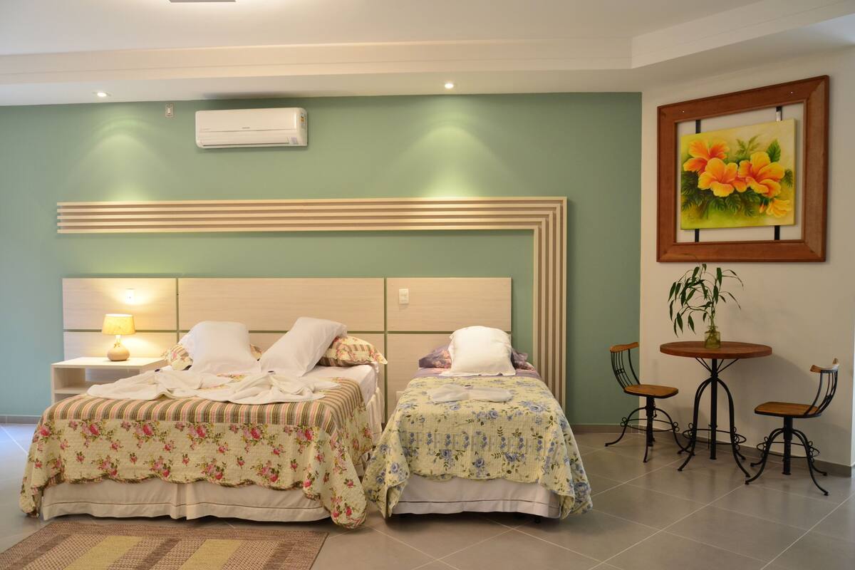Un dormitorio con dos camas, una mesita de noche con lámpara, una pequeña mesa redonda con sillas y un cuadro en la pared.