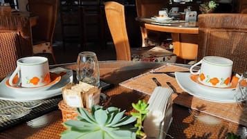 Una mesa de comedor dispuesta con platos blancos y tazas florales anaranjadas, rodeada de sillas y una mesa de mimbre.