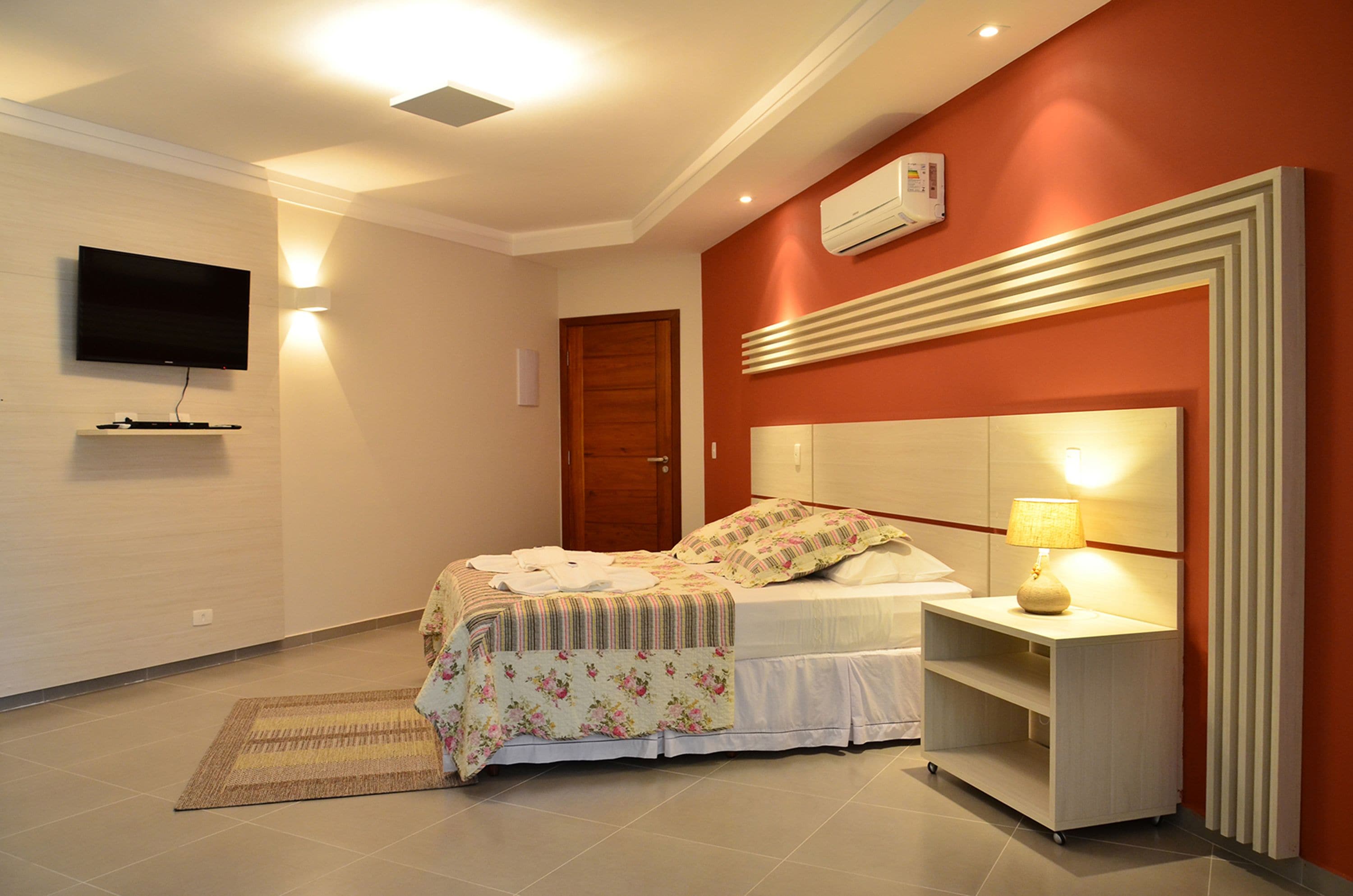 Quarto com cama, mesa de cabeceira, luminária, TV e ar-condicionado fixado na parede.