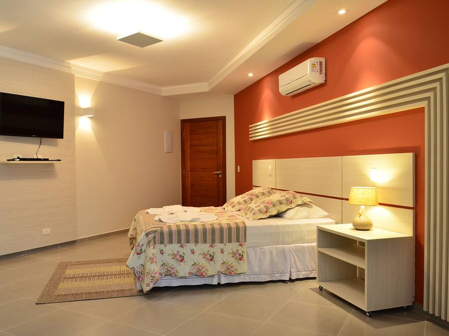 Quarto com cama, mesa de cabeceira, luminária, TV e ar-condicionado fixado na parede.