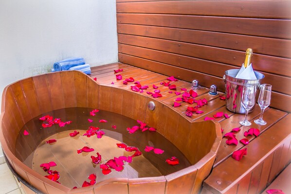 A wooden bathtub filled with water and rose petals, a bottle of champagne, and two glasses on a wooden bench. - Pousada L'essence (Paraty)