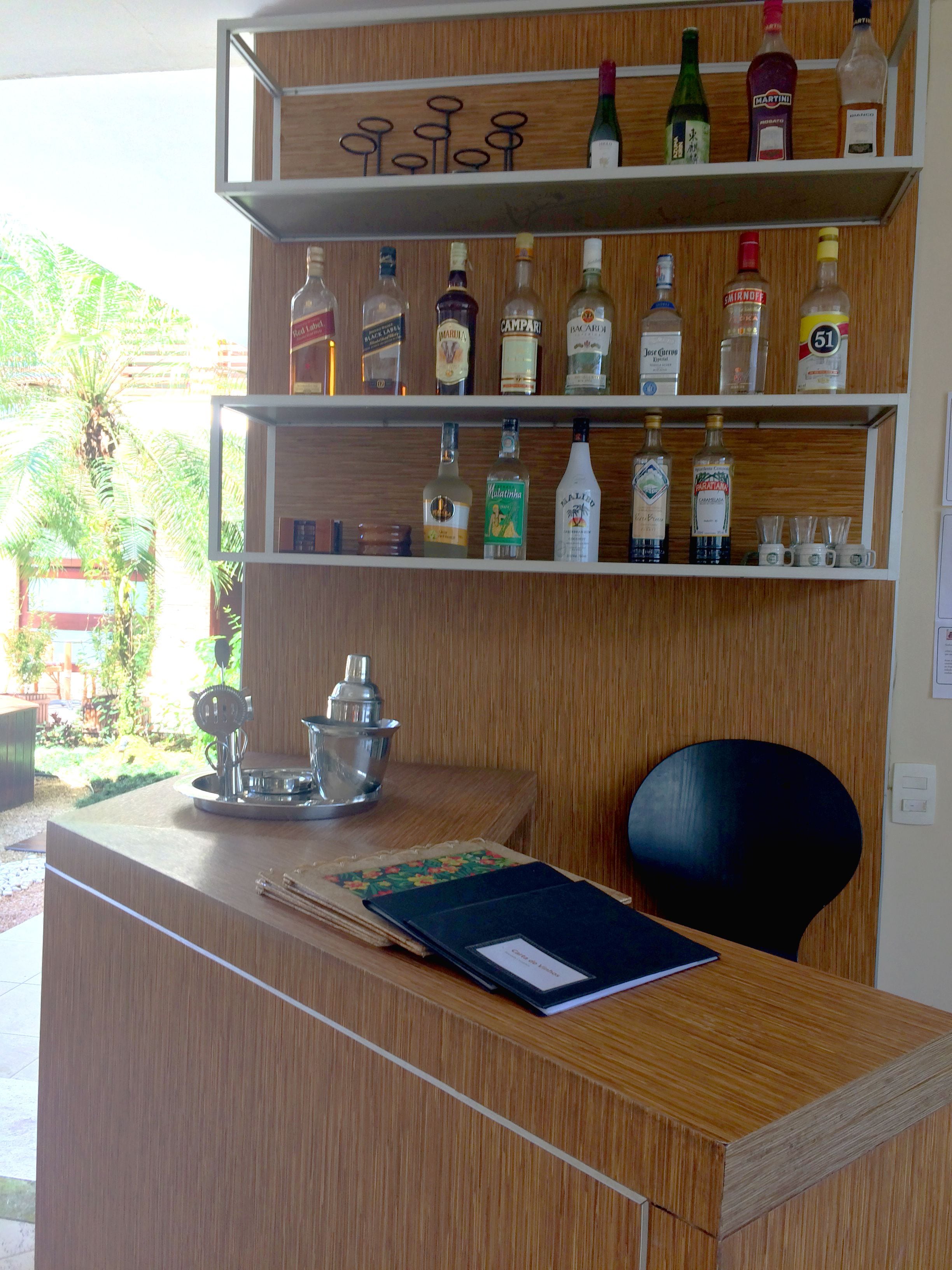 A reception desk with a wooden finish, a metal tray, and a notebook. Behind the desk, there are shelves with various liquor bottles and glasses.