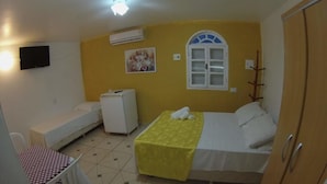 A room with a bed, a small table with a checkered tablecloth, a chair, a wardrobe, a window with a white frame, and an air conditioning unit on the wall. - Pousada Da Bia (RIO DAS OSTRAS)