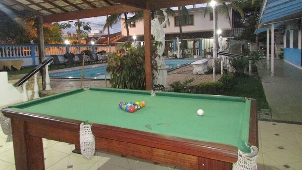 A pool table with balls, a covered area with a roof, a pool, and a building in the background. - Pousada Da Bia (RIO DAS OSTRAS)