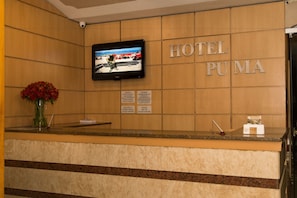 Ein Hotelrezeptionstresen mit einem Fernsehbildschirm, einer Blumenvase und einem Schild mit der Aufschrift 'HOTEL PUMA'.