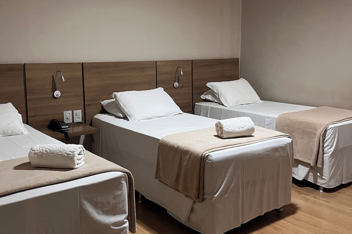 Une chambre d’hôtel avec trois lits, chacun recouvert d’une couvre-lit blanche et d’une couverture beige. On y trouve une tête de lit en bois, une table de chevet avec une lampe et un téléphone. La chambre a un plancher en bois et un luminaire au plafond.