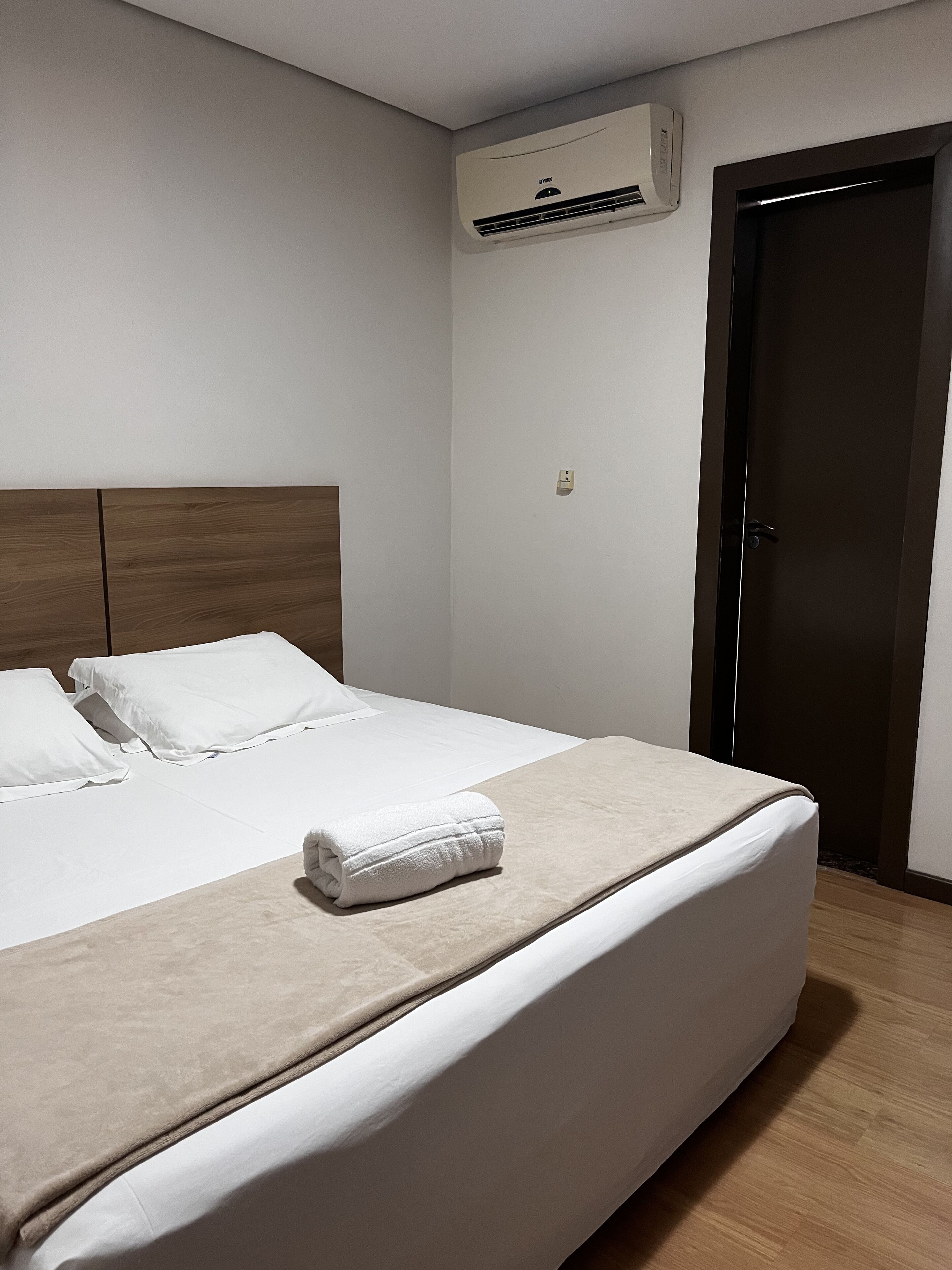 Un lit bien fait, avec du linge de lit blanc et une couverture beige, une tête de lit en bois, un climatiseur fixé au mur et une porte fermée.