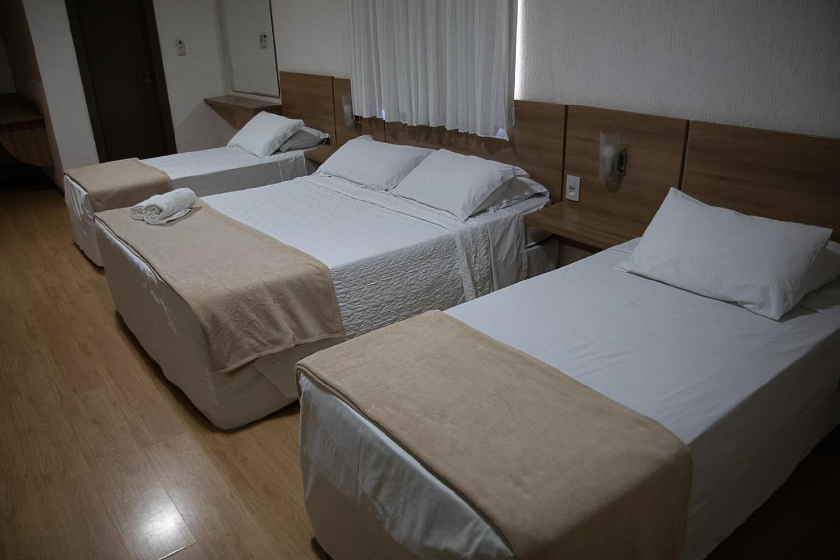 A hotel room with three single beds, each with a beige cover and white pillows. The room has wooden flooring and a wooden headboard.