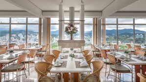 Comedor con mesas dispuestas para el servicio, amplias ventanas que ofrecen vista a la ciudad y a la montaña, y un arreglo floral central.
