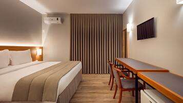 A hotel room with a bed, a desk, two chairs, a flat-screen TV, and an air conditioning unit.