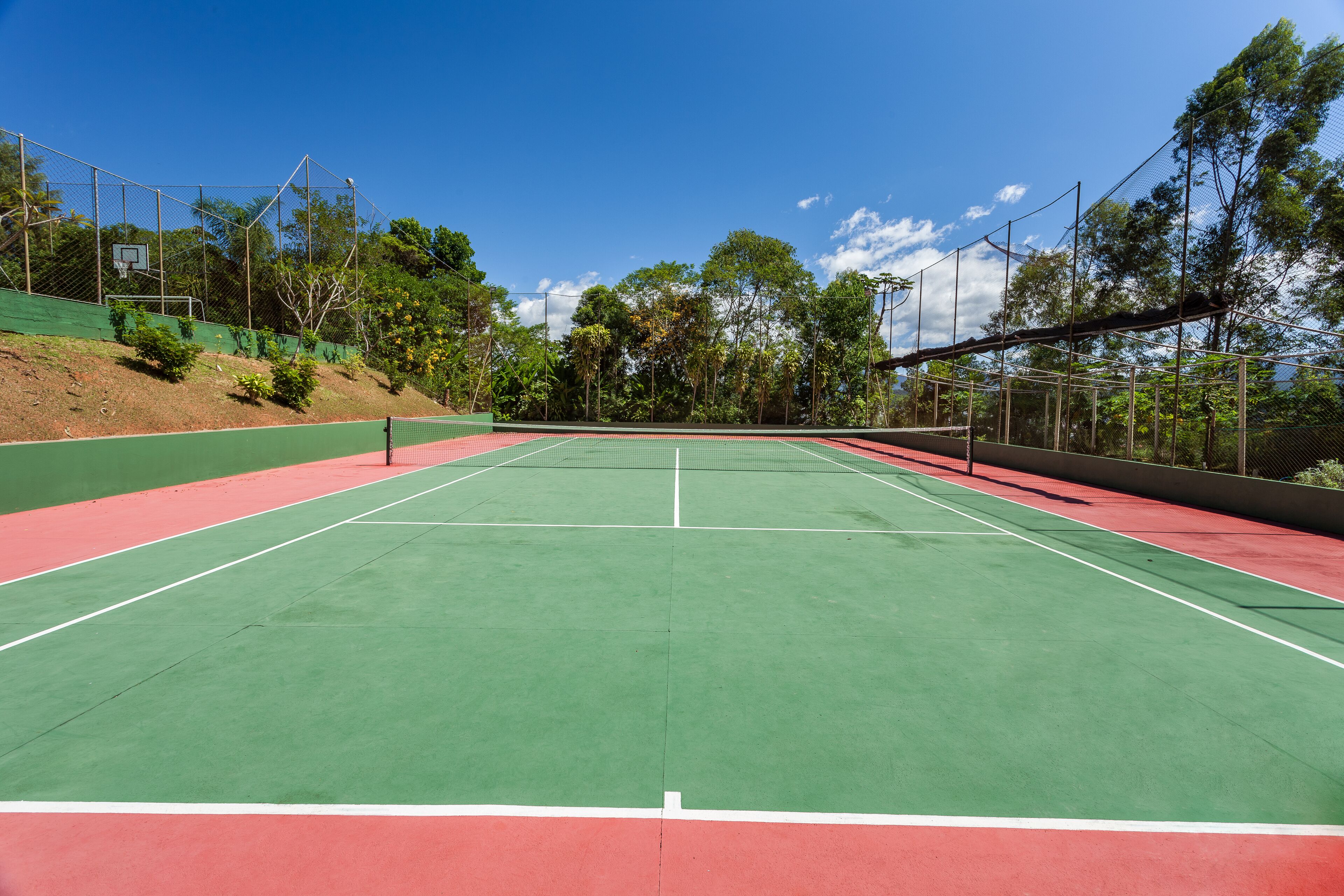 Una cancha de tenis con superficie verde y bordes rojos, rodeada de árboles y un cielo despejado.