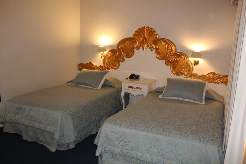 A hotel room with two beds, each with a patterned bedspread and pillows, a nightstand with a phone, and wall-mounted lamps.