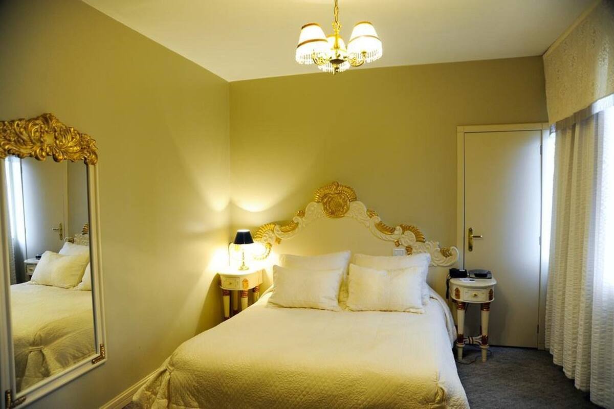 A hotel room with a large bed, a chandelier, a mirror, a bedside table, and a door.