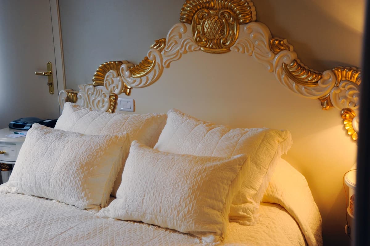 A bed with a white quilt and two pillows, a gold ornate headboard, and a bedside table with a lamp.