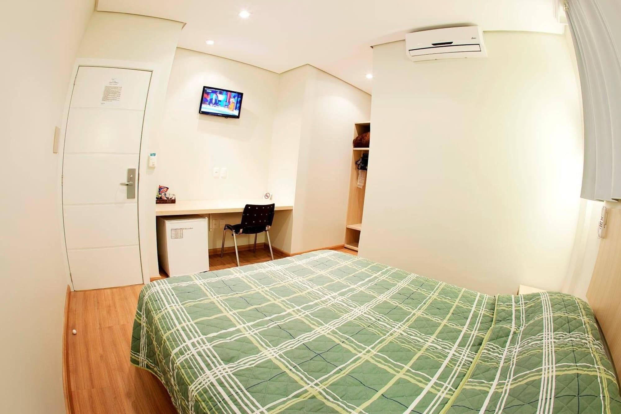 Un dormitorio pequeño y bien iluminado con cama, escritorio, silla y televisor montado en la pared.