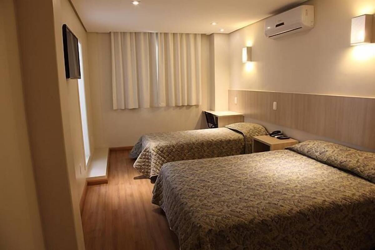 A hotel room with two beds, a bedside table with a lamp, and a window with curtains.