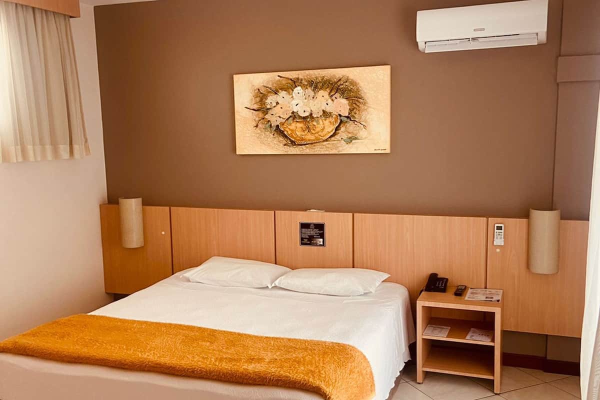 Une chambre d’hôtel avec un lit, une table de chevet avec un téléphone, un climatiseur fixé au mur et un tableau au-dessus du lit.