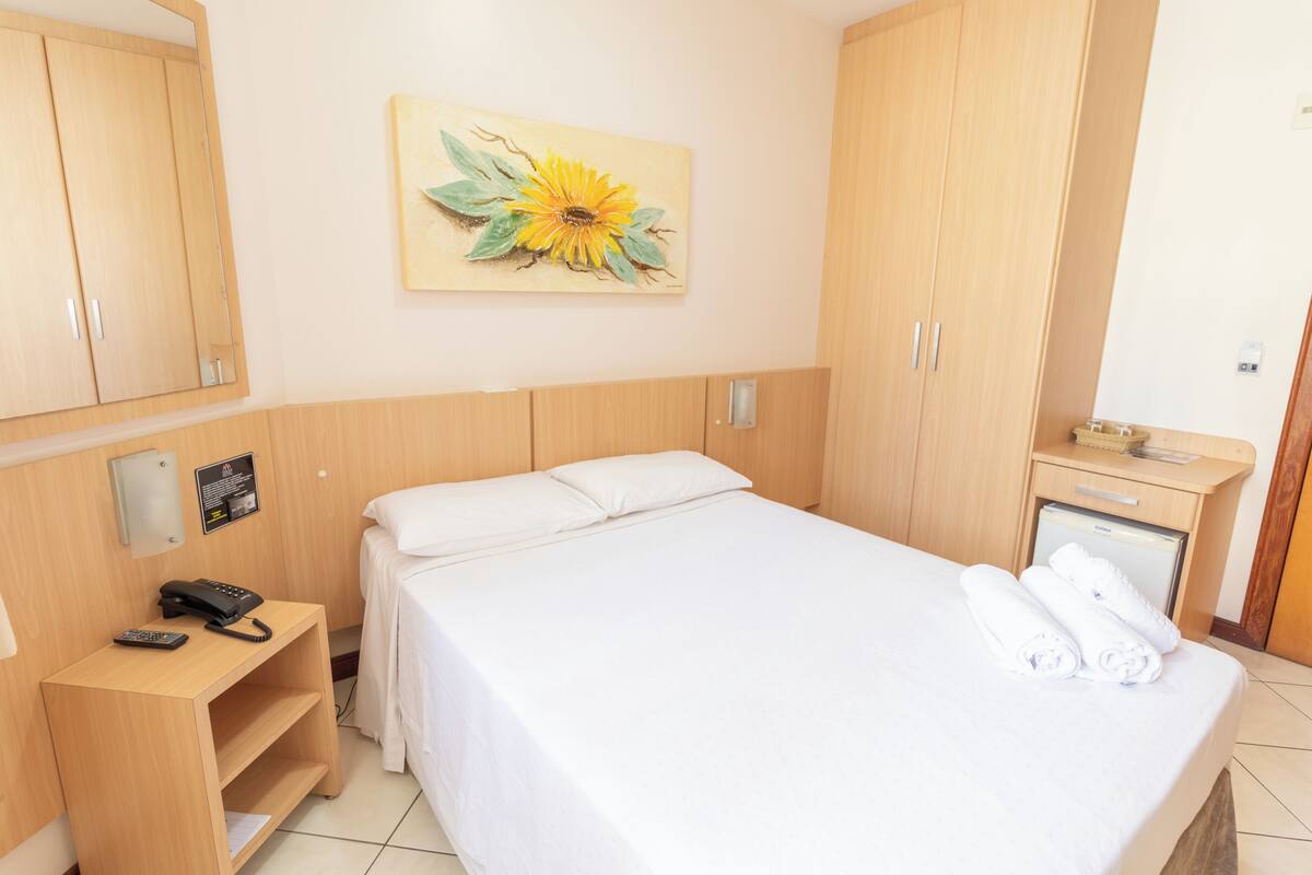 Une chambre d’hôtel avec un lit, un bureau avec un téléphone, une petite armoire et un tableau représentant un tournesol accroché au mur.