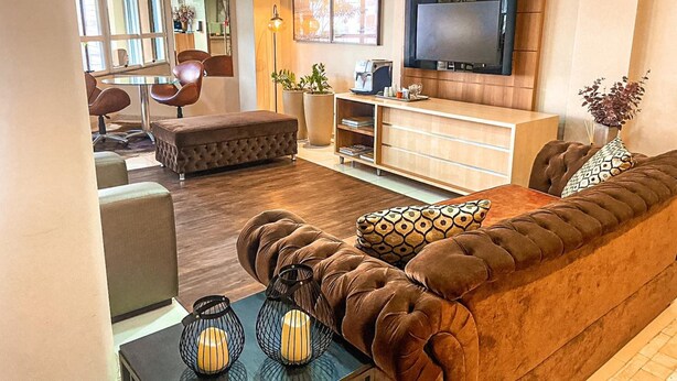 A modern living room with a brown sofa, a coffee table, and a TV mounted on the wall. - Diaudi Hotel (Sao Jose)