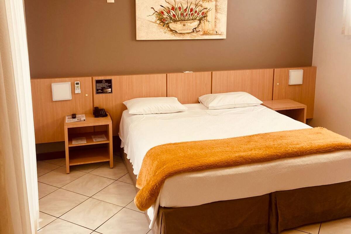 Une chambre d’hôtel comprenant un lit, une table de chevet, une commode, un climatiseur fixé au mur et un tableau.