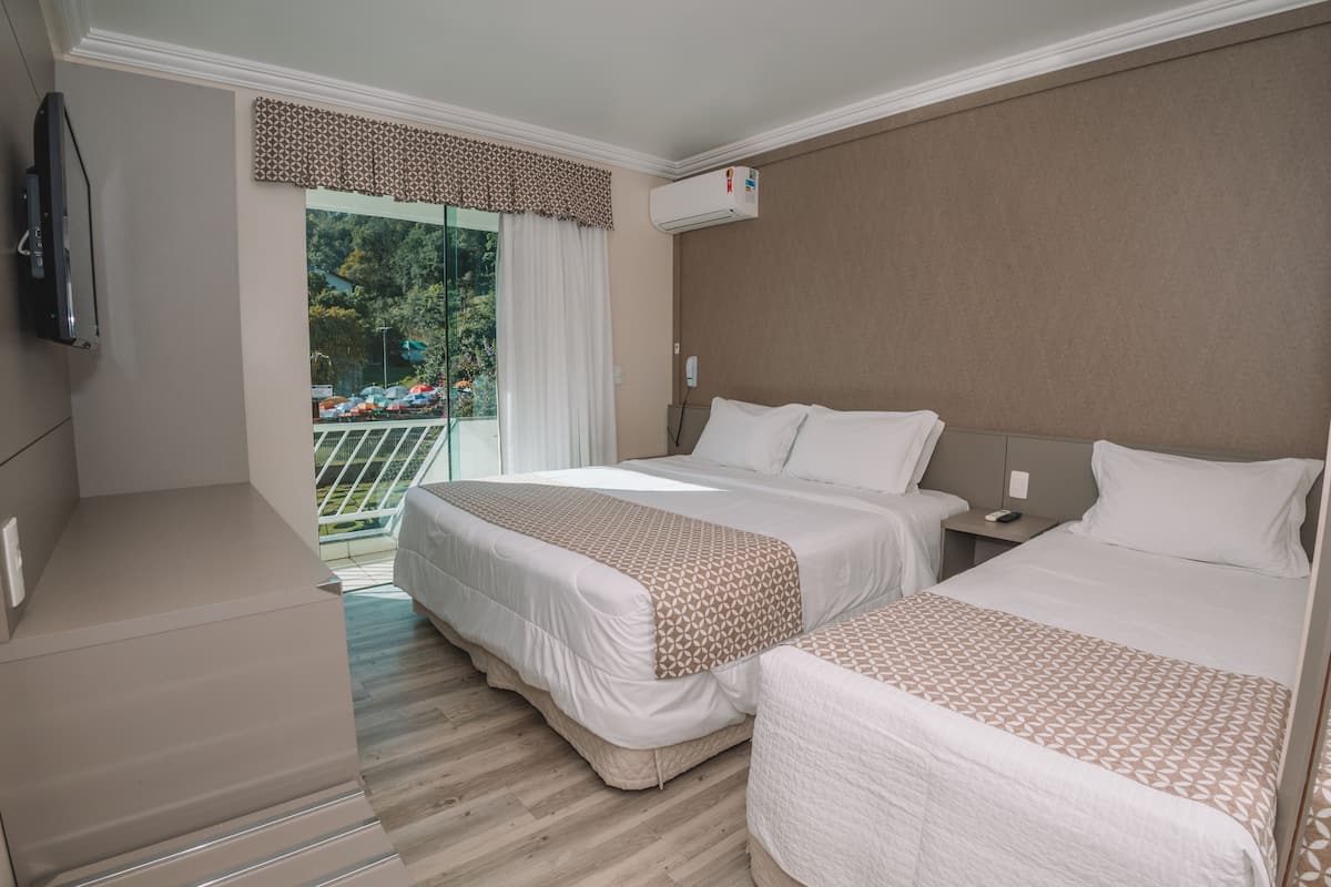 A hotel room with two beds, a TV, an air conditioner, and a balcony with a view of parked cars and greenery.