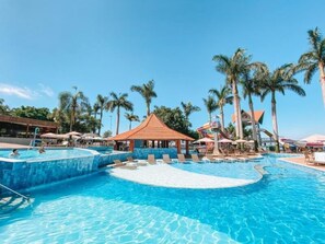 Una amplia piscina al aire libre con palmeras y un tobogán acuático, rodeada de tumbonas y sombrillas.