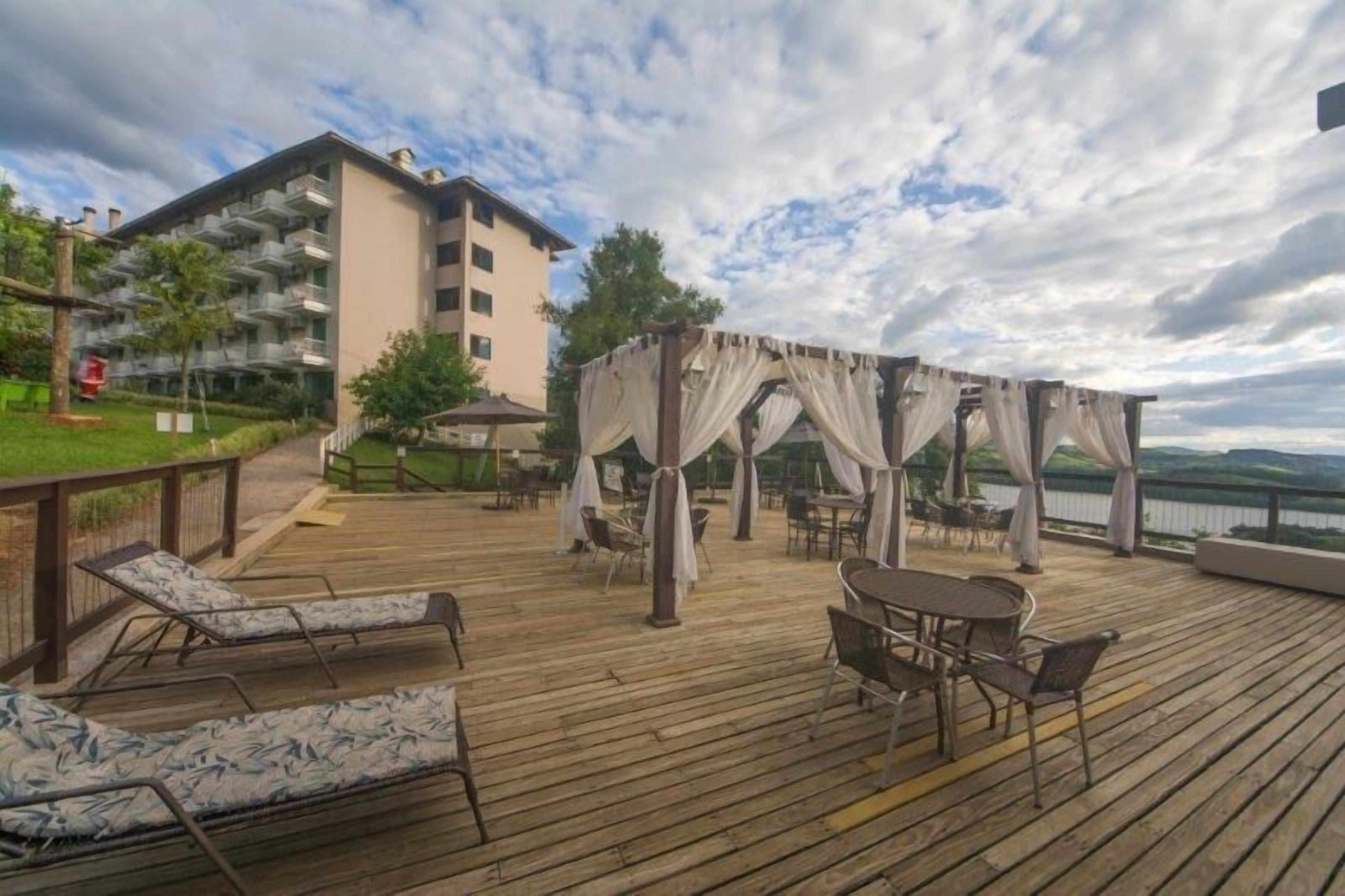 Terraza de madera con sillones de descanso y un área de estar cubierta, con vista a una masa de agua y un edificio con balcones.