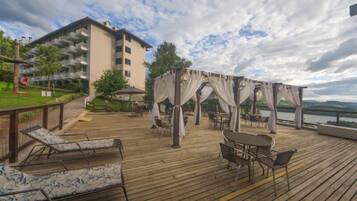 Terraza de madera con sillones de descanso y un área de estar cubierta, con vista a una masa de agua y un edificio con balcones.