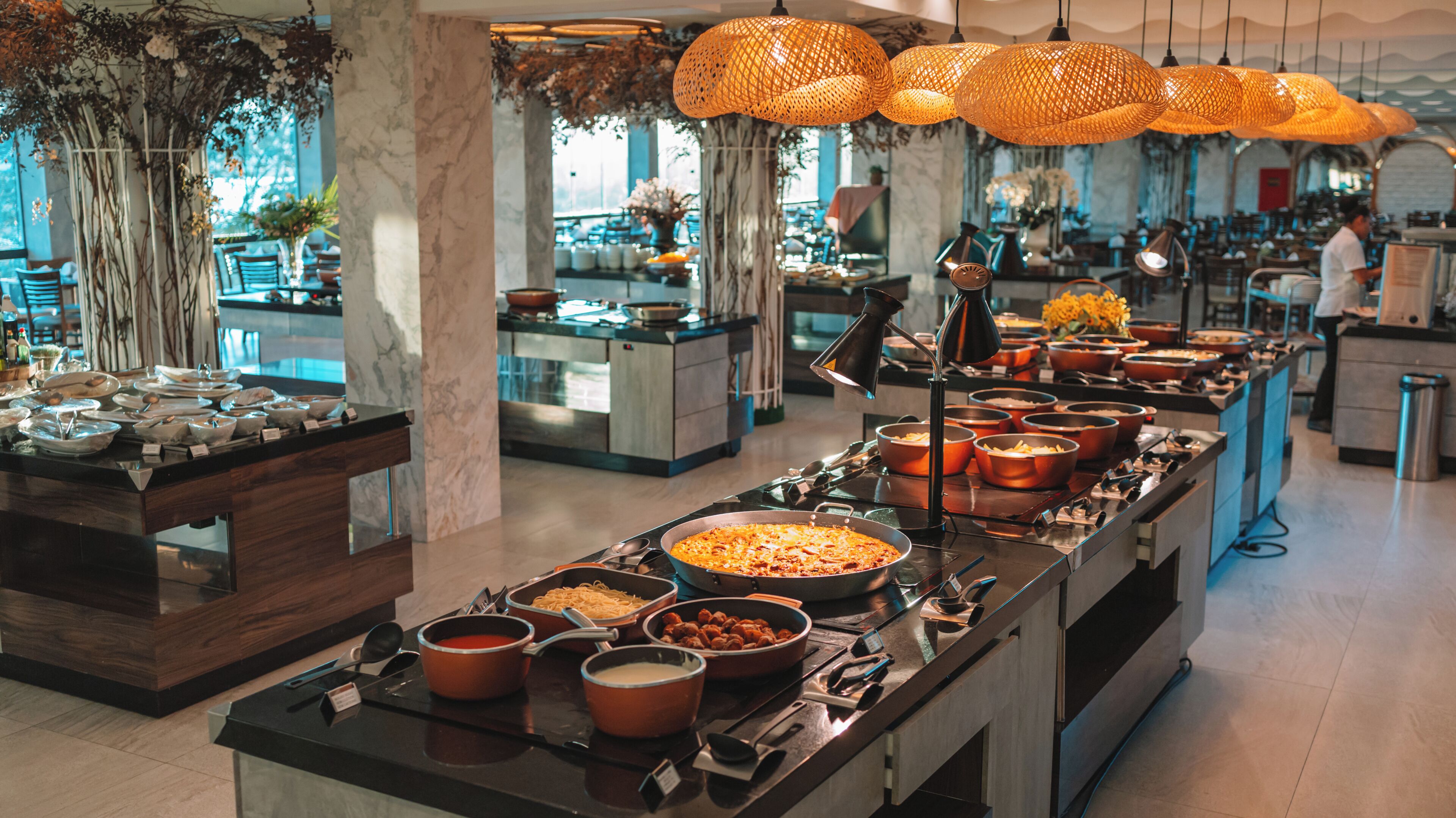 Disposição de buffet com diversos pratos, um pilar de mármore e luminárias pendentes alaranjadas.
