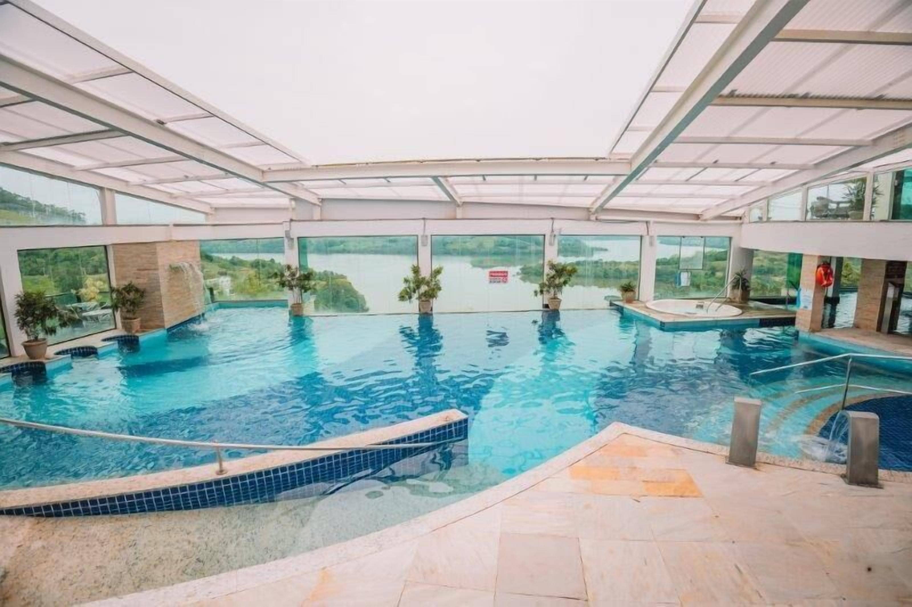 An indoor swimming pool with clear glass walls, potted plants, and a view of the outdoors.