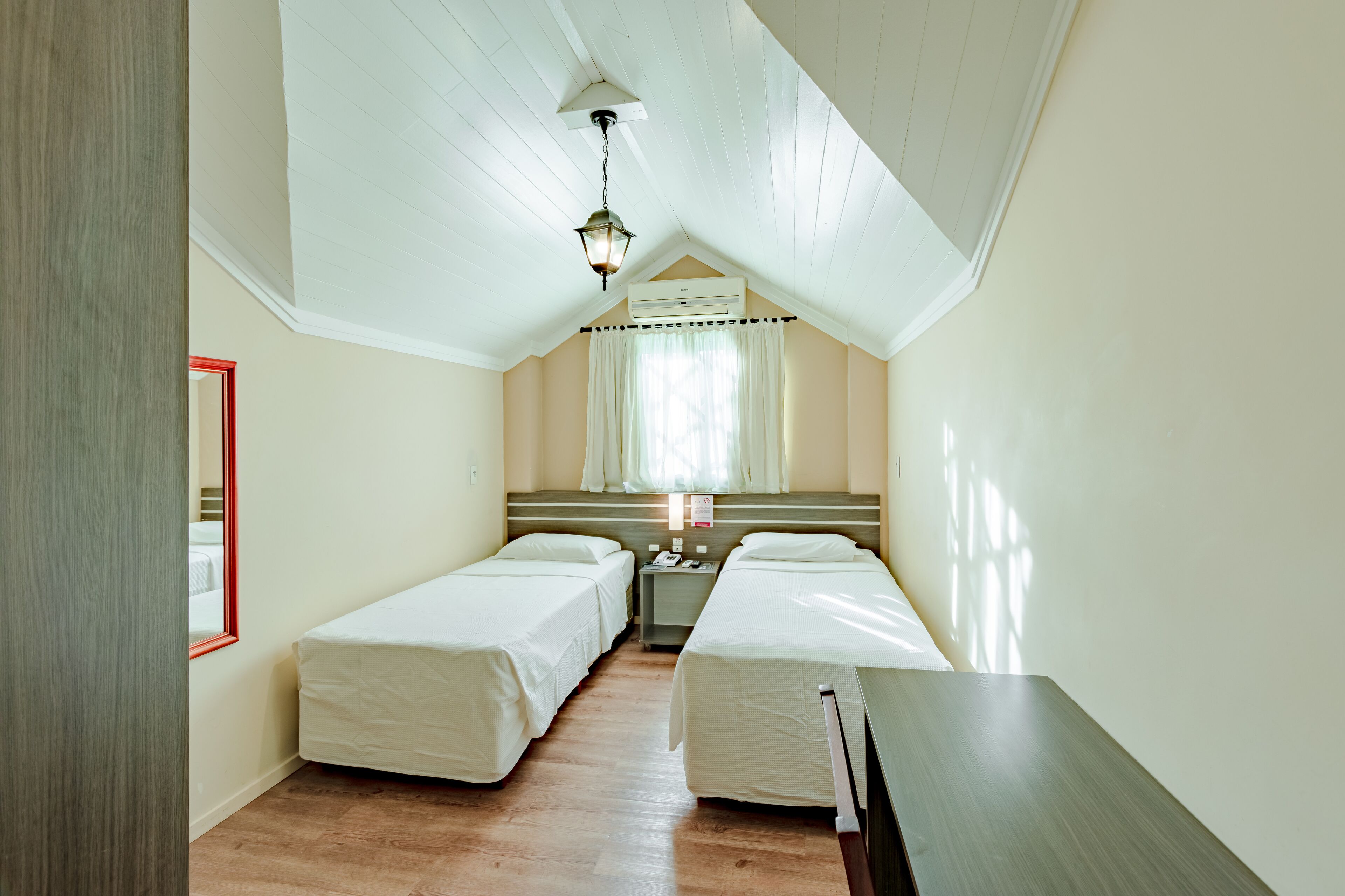 A small, single-room hotel room with two single beds, a wooden floor, a ceiling fan, and a window with curtains.