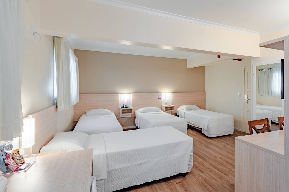 Ein Hotelzimmer mit vier Betten, einem Schreibtisch und einem Stuhl. An den Betten stehen Nachttische mit Lampen, und an der Wand befindet sich ein Spiegel.