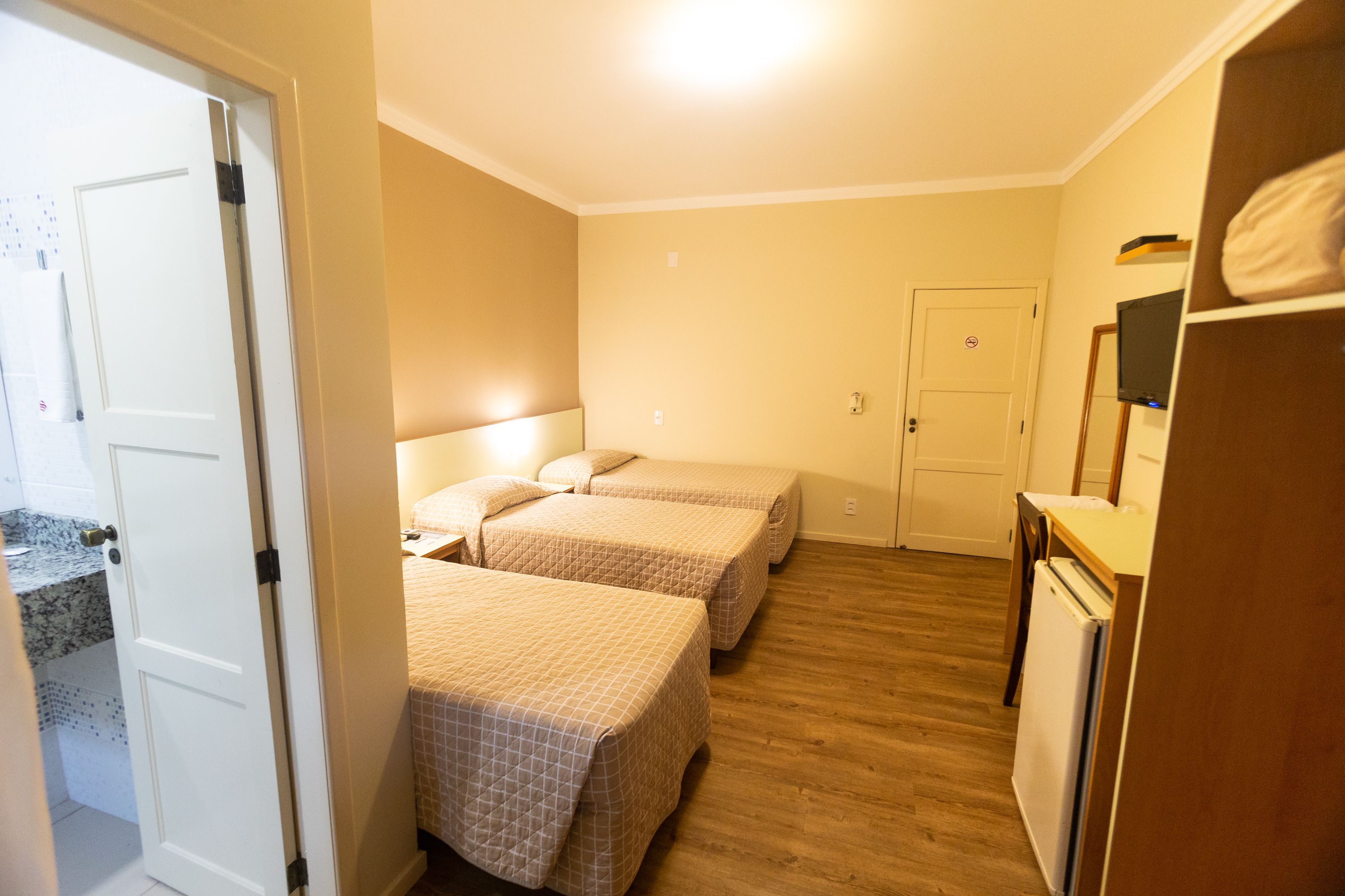 Una habitación pequeña y luminosa con dos camas, piso de madera, puerta blanca y una pequeña zona de kitchenette.